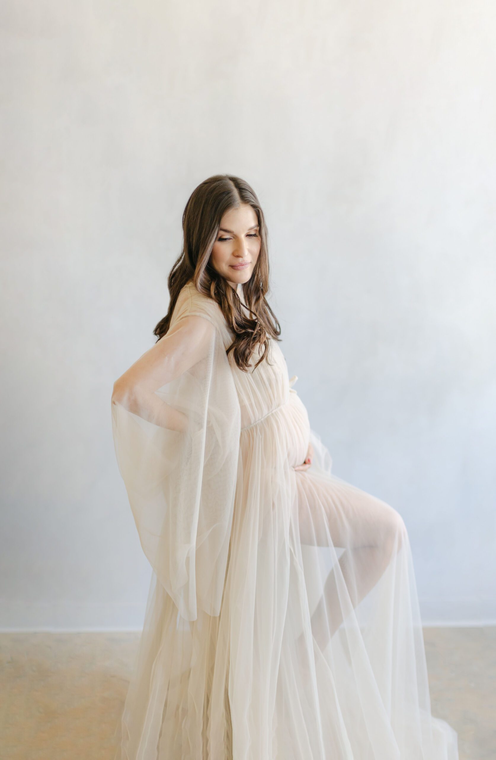 expectant long hair brunette in tulle gown posing on stool